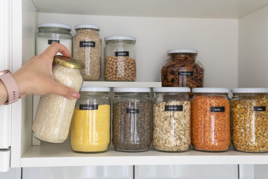 Hand picking a jar of rice from an organized pantry shelf with labelled jars of grains, pulses, and spices.