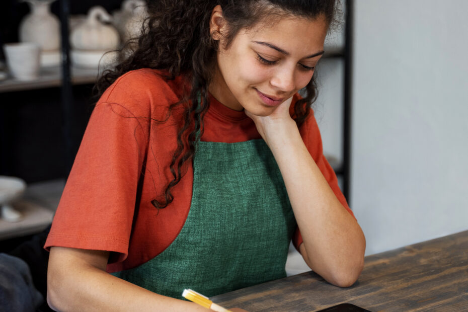 Indian woman in a casual green apron writing a weekly meal plan at a wooden table with her phone beside her — representing mindful kitchen planning.