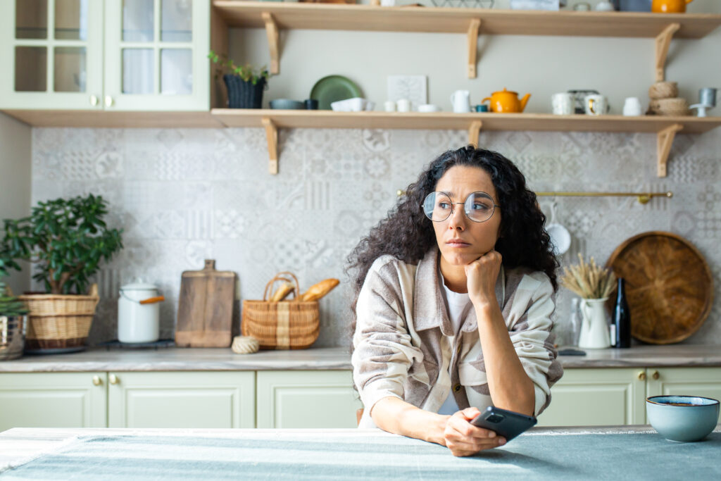 Indian woman sitting thoughtfully in a modern kitchen, holding a phone, looking concerned — representing daily meal planning stress before finding a solution. Aara Living