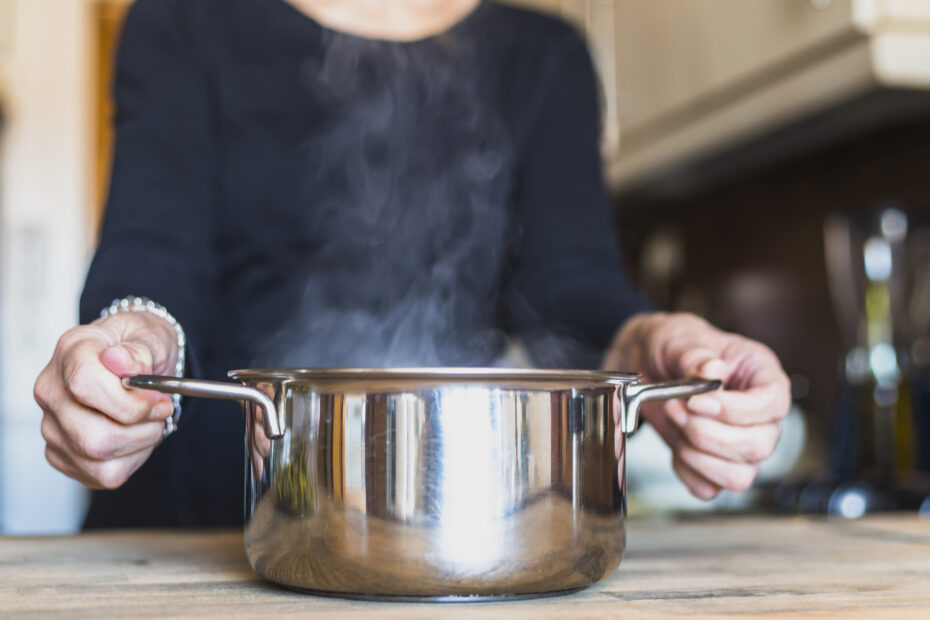 Woman holding a steaming pot of freshly cooked rice in a home kitchen, representing traditional open-pot rice cooking methods in India.