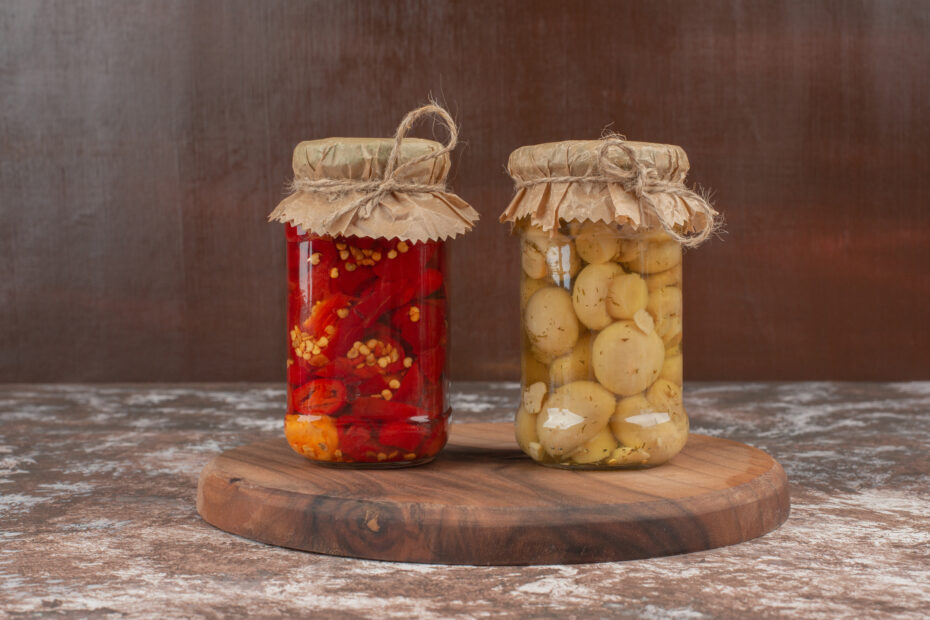 Homemade fermented vegetables in glass jars — red peppers and mushrooms preserved in brine — placed on a rustic wooden plate. Aara Living