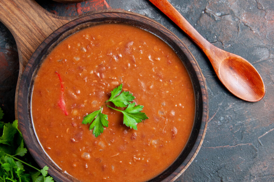 Bowl of hot rajma curry garnished with coriander on a rustic red background with garlic and raw kidney beans on the side. Aara Living.